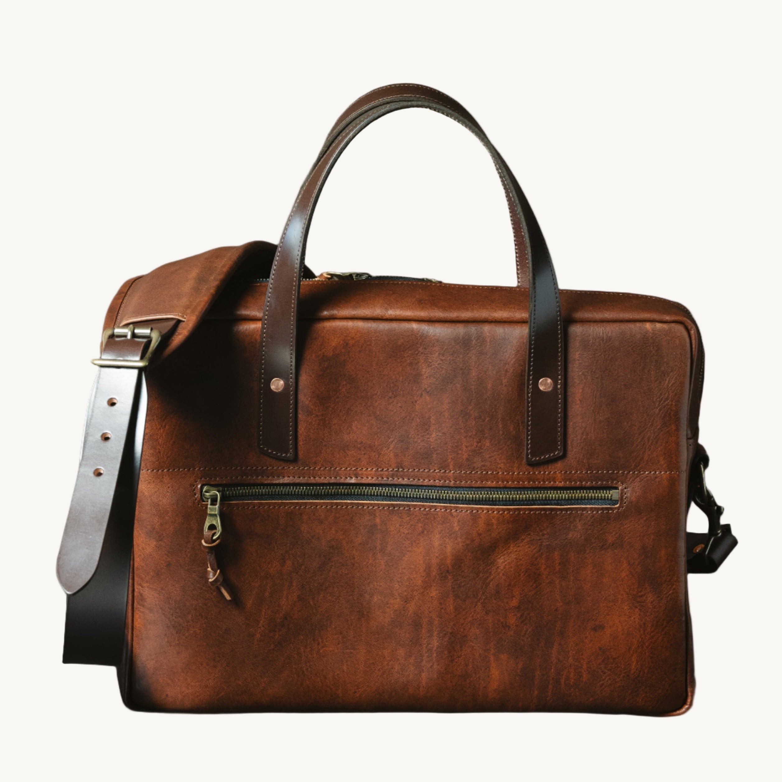 A brown leather briefcase with a cognac color scheme, featuring a shoulder strap, handle, and zipper detailing against a dark background.