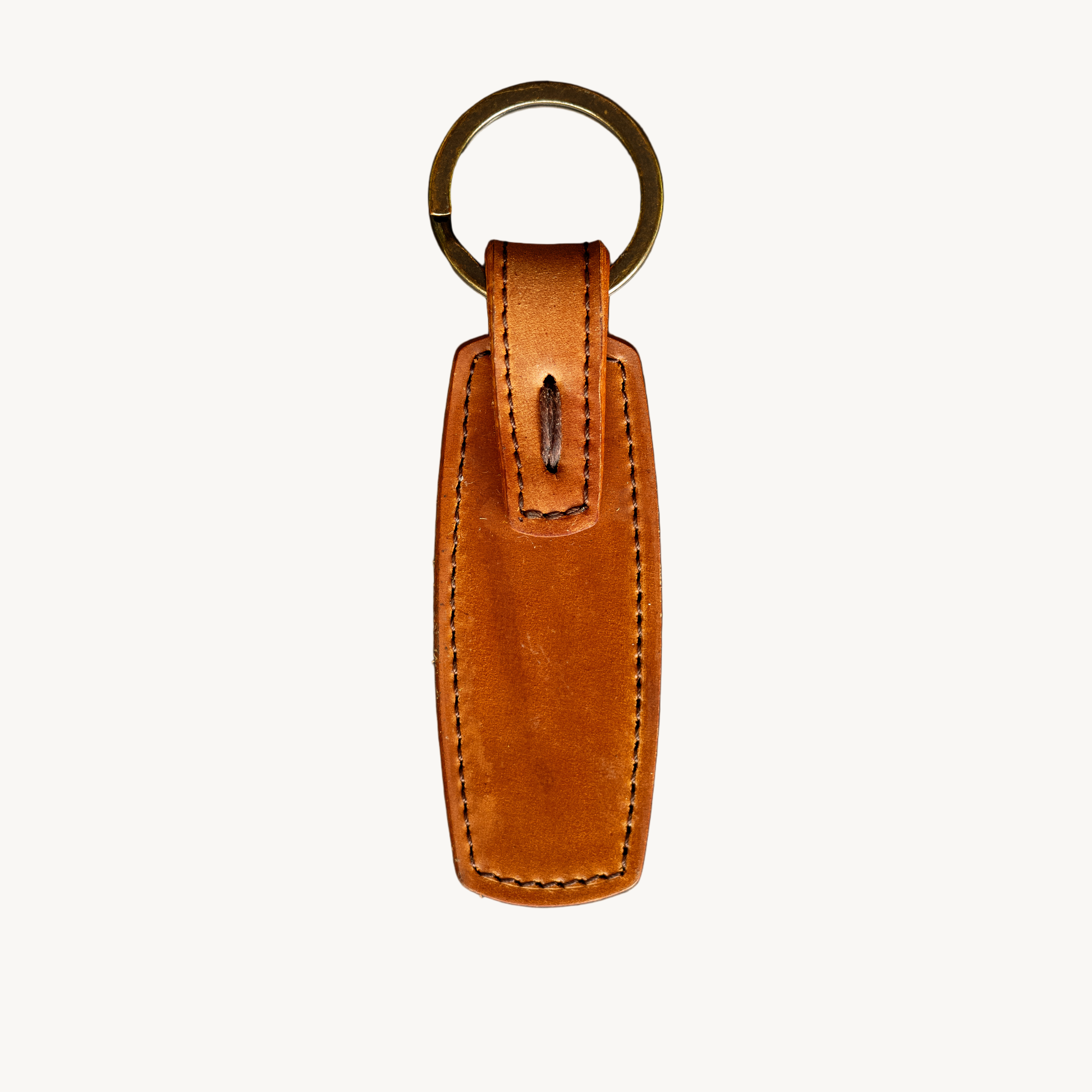 A brown leather key ring with a metal antique brass keyring and hand-stitched detail, laid on a textured surface.