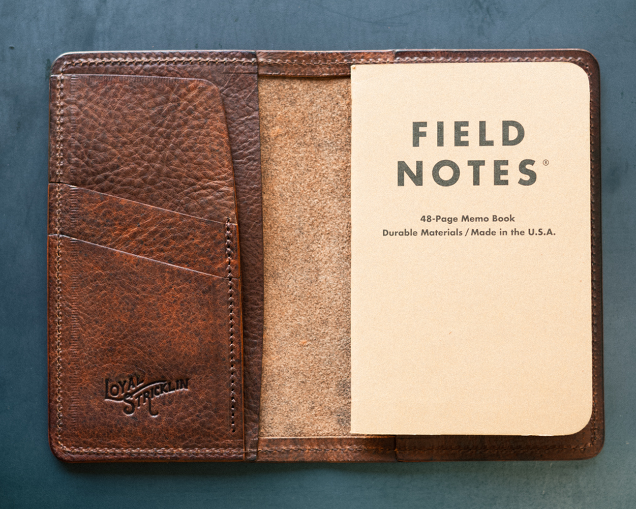 A brown leather wallet opened to show the interior, which includes a Field Notes notebook, card pockets, and a space for a passport or notebook.