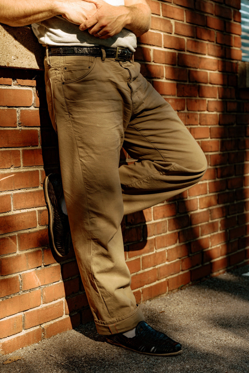 Person leaning against a brick wall with a guitar, wearing beige pants and a white shirt.